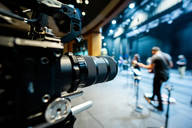 Closeup of professional video camera with lens recording unrecognizable people standing on stage illuminated by lights in studio. Youtube channel sponsorship: a grounded overview